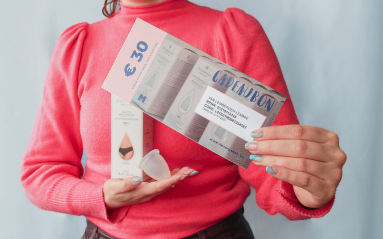 Beeld: vrouw houdt cadeaubon van LM Oost-Vlaanderen voor herbrbuikbaar menstruatiemateriaal vast, met een menstruatiecup en in verpakking menstruatieondergoed in de andere hand.