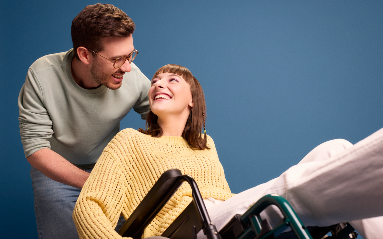 Koppel kijkt liefdevol naar elkaar, jonge vrouw zit in een rolstoel en wordt gekanteld door haar vriend