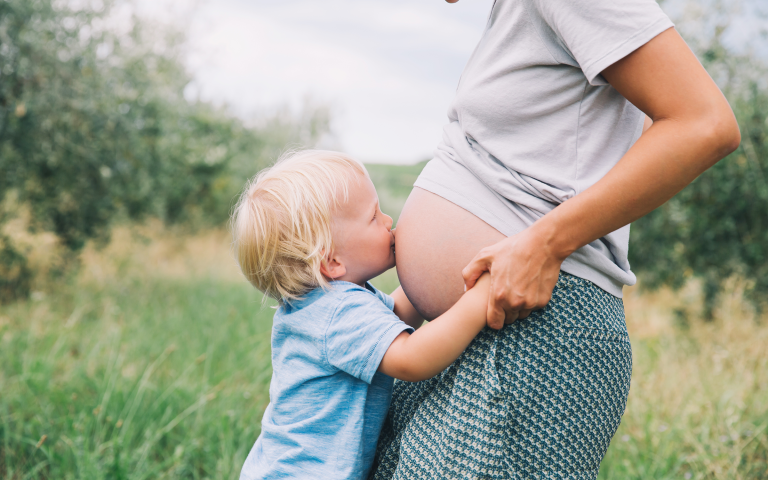 zwangere vrouw staat in de natuur met haar blote zwangere buik, blond kleutertje geeft een kus op de buik