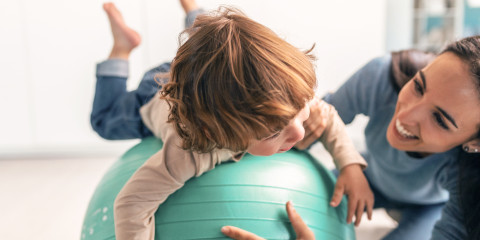 Ouder-kleuterturnen: kleuter probeert liggend op zijn buik te balanceren op een sportbal, mama lacht zoon toe en helpt hem met het balanceren