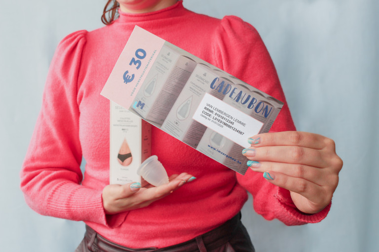 Beeld: vrouw houdt cadeaubon van LM Oost-Vlaanderen voor herbrbuikbaar menstruatiemateriaal vast, met een menstruatiecup en in verpakking menstruatieondergoed in de andere hand.